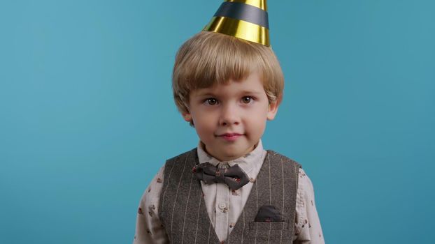 Smiling little birthday boy in cone hat on blue wall background. Happy emotional child. Amazement in eyes, happy childhood