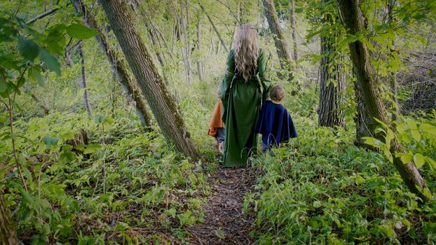 Two little toddler boys cosplay gnomes or hobbits with elf woman walking in green forest. Unrecognizable fairy tale characters. Halloween, kids concept. Amazing light.