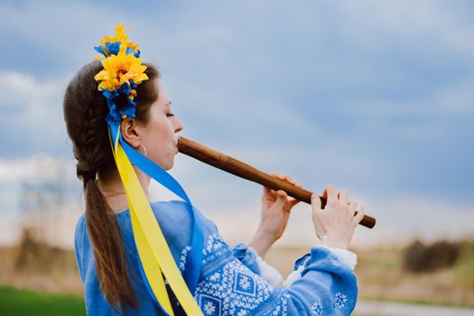 Woman playing woodwind wooden flute - ukrainian sopilka outdoors. Folk music, culture concept. Musical instrument. Lady in traditional embroidered shirt - blue vyshyvanka.
