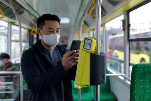 Asian man in public transport bus in protective medical mask buys e-ticket by phone, passenger in empty bus