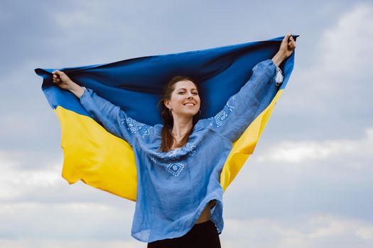 Happy free ukrainian woman with national flag on dramatic sky background. Portrait of lady in blue embroidery vyshyvanka shirt. Ukraine, independence, patriot symbol