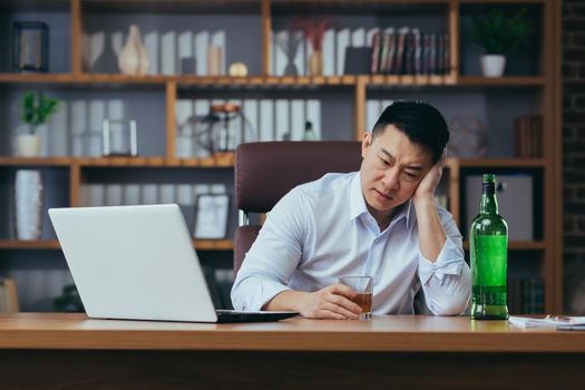 Asian businessman fails, man in despair drinks hard liquor, sitting late at the table in the office