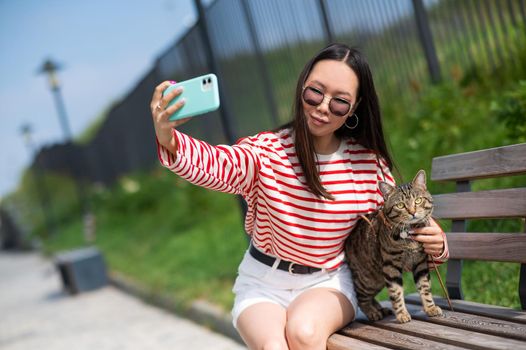 Young woman sits on a bench with a tabby cat and takes a selfie on a smartphone outdoors.