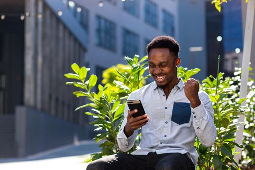 happy african american man in casual clothes sitting on bench outdoors with mobile phone