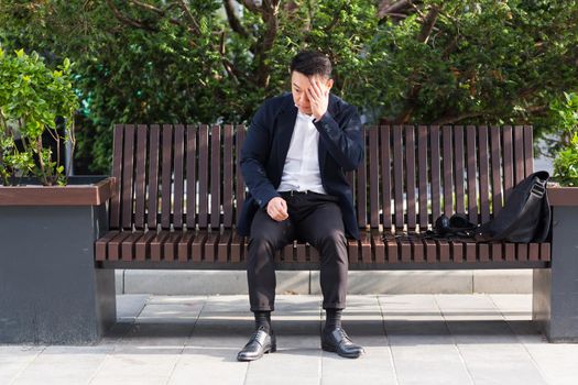 exhausted young asian business man office worker sitting on bench in city park outdoors