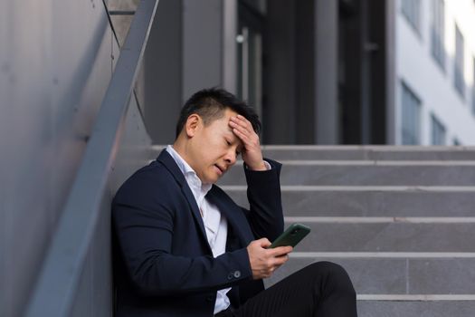 Asian businessman broker reports bad news talking on the phone, sitting near office