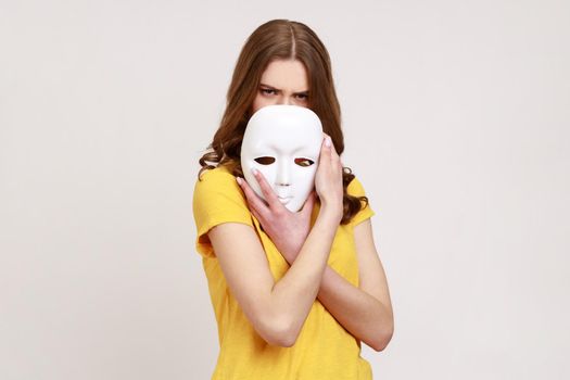Portrait of teenager girl with brown hair in yellow casual style T-shirt covering half of face with white mask, multiple personality disorder.