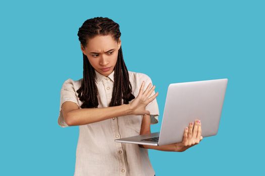 Woman with dreadlocks shows stop gesture to laptop display, forbidden content, looking with disgust