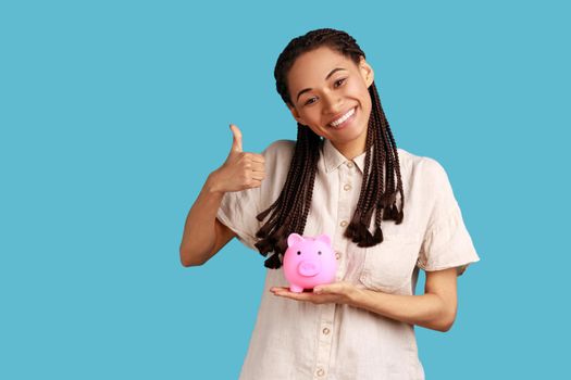 Pleased woman with dreadlocks holding pig money box in hands, saving, showing thumb up.