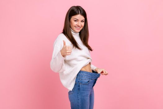 Side view portrait of happy beautiful woman showing thumb up and her slim waist and big trousers.