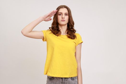 Yes sir. Young patriotic girl in casual yellow T-shirt holding hand near head, saluting, honor and glory, looking seriously at camera.