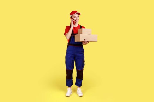 Delivery woman standing with two parcels in hands, talking mobile phone with client.