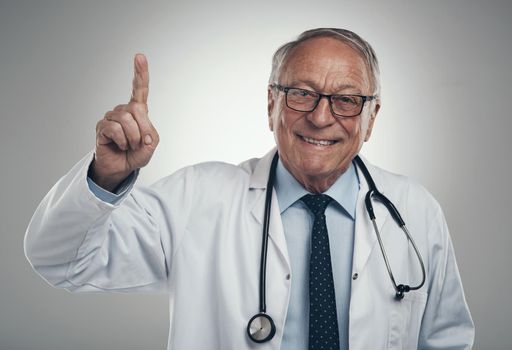 Ah yes Ive got a great idea. an elderly male doctor holding his finger up in a studio against a grey background.