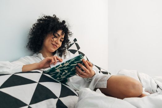 Happy young mixed race woman relaxes reading journal notes on bed. Copy space.