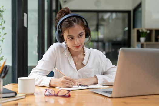 Online education, e-learning. young woman studying remotely, using a laptop, listening to online lecture, taking notes while sitting at home, smiles