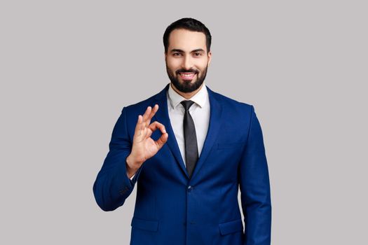 Bearded man standing, looking at camera showing Ok sign gesture, expressing positive emotions.