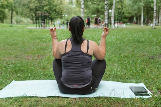 A charming brunette woman plus-size body positive practices sports in nature. Woman does yoga in the park on a sports mat