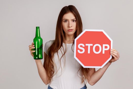 Anxious beautiful woman showing alcoholic beverage beer bottle and stop sign, warning and worrying.