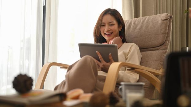 Happy young woman resting on armchair and watching videos, surfing internet on digital tablet