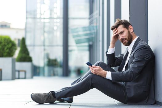 Office worker sitting near office outdoors got bad news, depressed and tired reads bad news