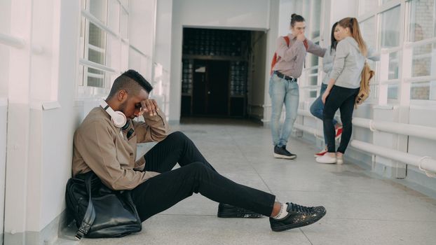 View of upset serious african-american male student sitting on floor in white spacious university corridorwhile his groupmates are standing communicating