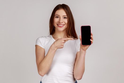 Happy woman pointing at cellphone, advertising gadget or online application, cellular tariffs plan.