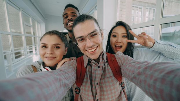 Point of view of four positive cheerful attractive multi-ethnic friends talking selfie photos holding smartphone and having fun while standing in corridor of university