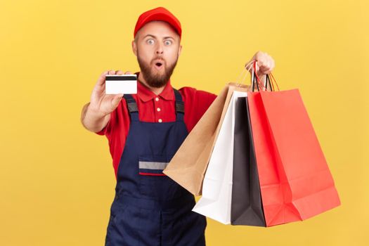 Shocked deliveryman in blue uniform holding shopping bag and credit card, cashless payments.