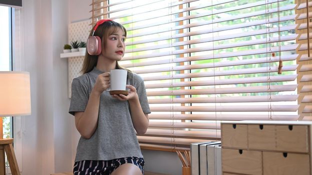 Satisfied young woman holding coffee cup and looking through the window, enjoy stress free peaceful mood