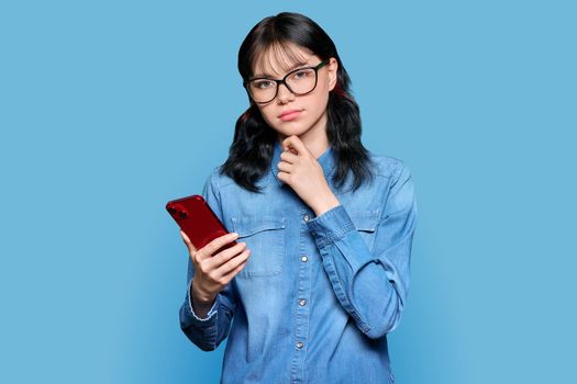 Serious teenage girl holding smartphone looking at camera, on blue background