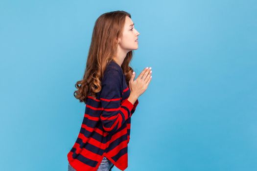 Please, need your help. Side view portrait of woman wearing striped casual style sweater holding hands in praying gesture, pleading with desperate face.