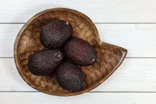Four whole ripe brown avocados in wooden carved bowl on white boards desk. Tabletop view.