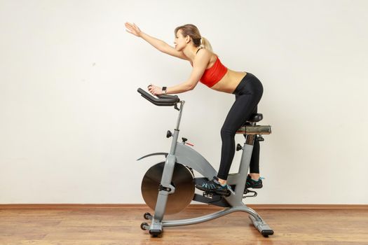 Slim woman riding exercise bike and raised arm, doing sport exercises during cardio workout.