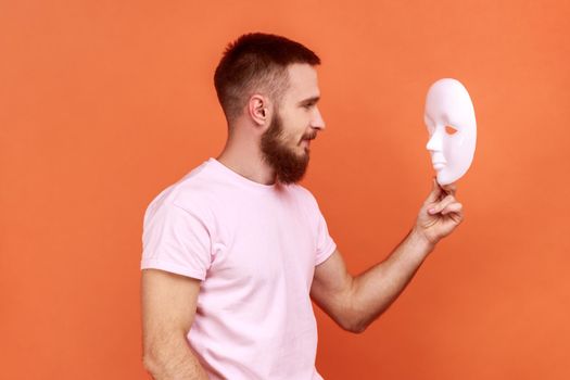 Man looking at white mask in hands with attentive look, trying to understand hiding personality.