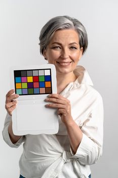 Mature woman holds color checker smiling at camera. Gray haired woman in white blouse shows target for color adjustment. Isolated on white background