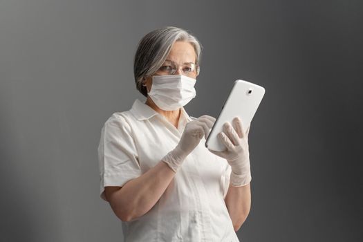 Serious female doctor works with tablet computer. Caucasian woman in white protective uniform uses digital tablet looking at camera while standing on gray wall background.