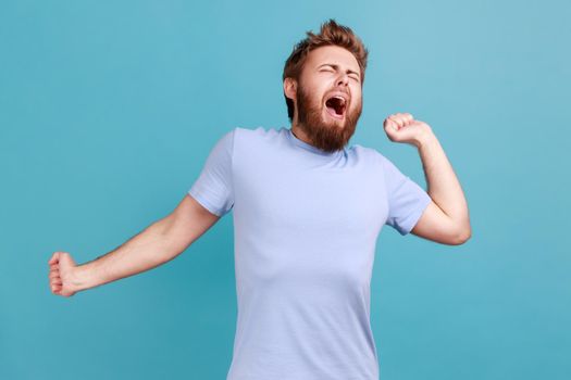 Man in T-shirt standing and yawning with closed eyes and raised arms feels sleepy, needs more energy
