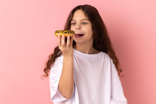 Delighted cute little girl licking delicious donut, looking with desire to eat sweet dessert.