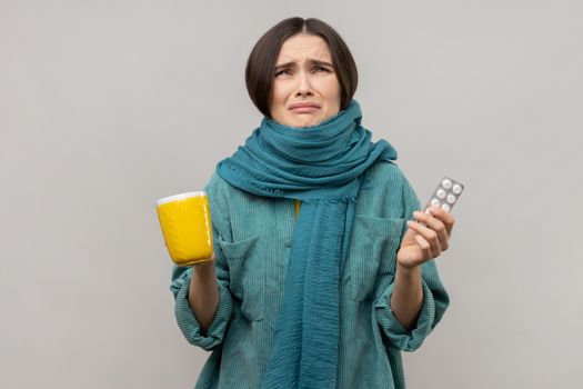 Irritated young woman wrapped in scarf holding tea, napkin, feeling despair about influenza.