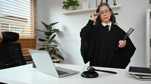 Positive mature female judge, lawyer or attorney in robe gown uniform standing at her personal office. Law and justice concept