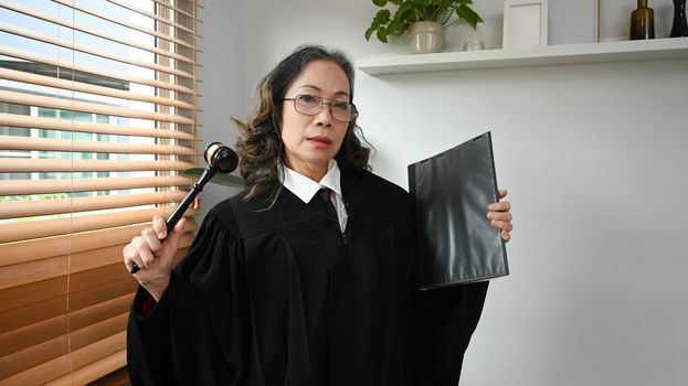 Confident mature female judge, lawyer or attorney in robe gown uniform with gavel hammer mallet. Law and justice concept
