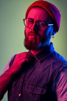 Hipster man in glasses, looking at camera, holding hand on his collar, wearing red beanie hat.