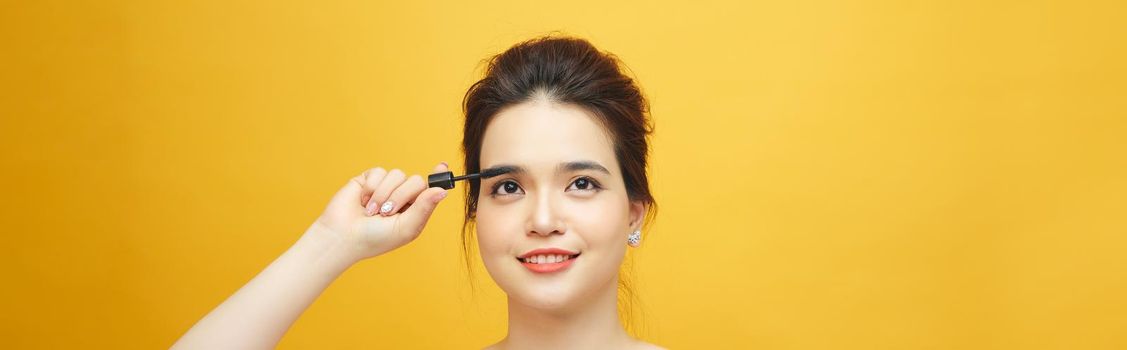 Closeup portrait of attractive young woman putting some mascara onto her eyelashes with make up brush over yellow background