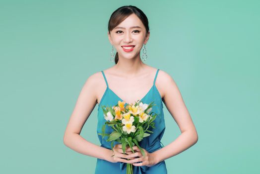Attractive Asian woman in blue dress holding bouquet of flowers over blue background