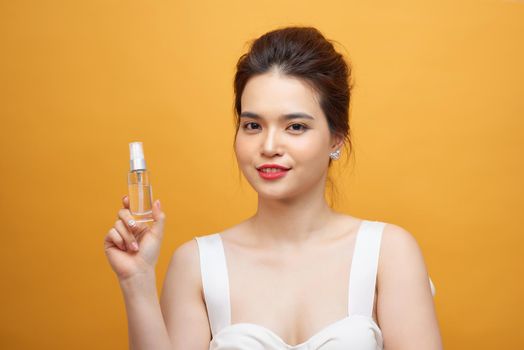 Beautiful woman holding spray bottle on face isolated on yellow background 
