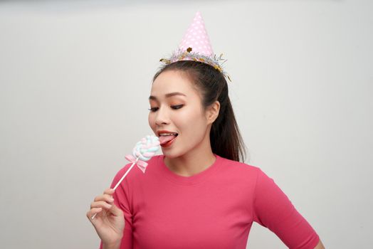 Blinking young woman in birthday hat holding, licking round lollipop celebrating 
