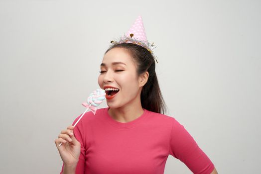 Crazy young woman in birthday hat biting round lollipop celebrating on white background.