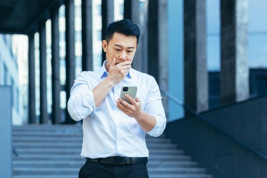 Upset Asian businessman reads news from phone screen, man disappointed with bad news