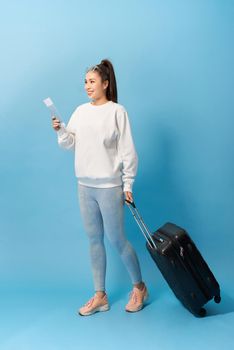 Portrait of trendy young girl standing with suitcase and holding passport with tickets, over blue background