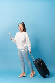 Portrait of trendy young girl standing with suitcase and holding passport with tickets, over blue background